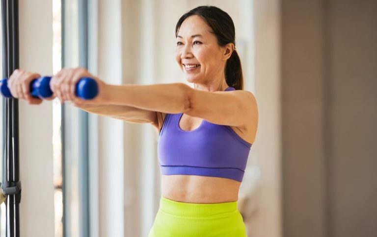 General Health Promotion & Fitness Optimisation A woman smiles while exercising with blue dumbbells in a bright, spacious room.